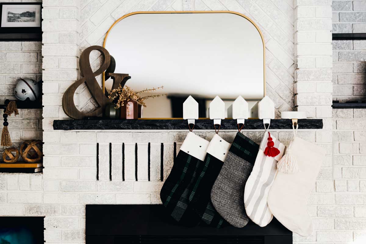 Camino moderno con mattoni bianchi, specchio dorato, mensola decorata e calze natalizie in colori neutri, dettagli in legno e tessuti, atmosfera invernale elegante.