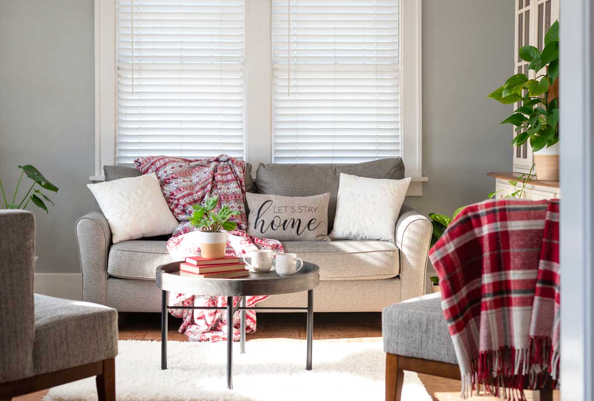 Soggiorno luminoso con divano grigio chiaro, cuscini neutri, plaid rosso, tavolino rotondo con pianta e libri, dettagli naturali e luce invernale che entra dalla finestra.