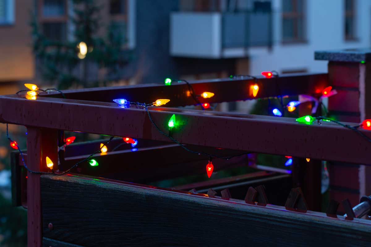 Dettaglio di una ringhiera da balcone decorata con una catena di luci natalizie multicolore. Le lampadine a led, di forma classica e colori vivaci (giallo, rosso, verde, blu), creano un’atmosfera allegra su uno sfondo urbano al tramonto.