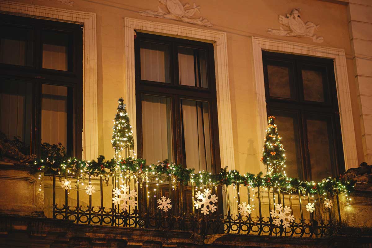 Balcone decorato per Natale con luci calde, ghirlande verdi e due piccoli alberi luminosi agli angoli. Fiocchi di neve bianchi applicati sulla ringhiera in ferro battuto. L’atmosfera è elegante e festosa, su una facciata storica con grandi finestre scure.
