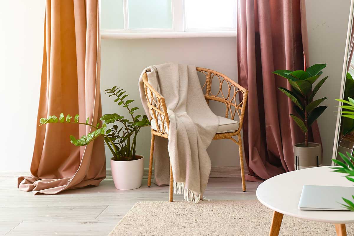 Soggiorno luminoso con tende lunghe color cipria, piante verdi, specchio e arredi in legno chiaro. Atmosfera accogliente e dettagli naturali.